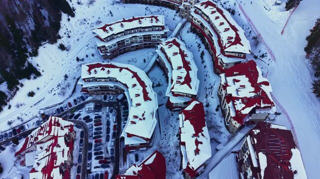 Snow blankets the rooftops of several buildings in a mountain resort. Cars are parked in organized rows in a snowy area. The scene shows a winding structure with many units.