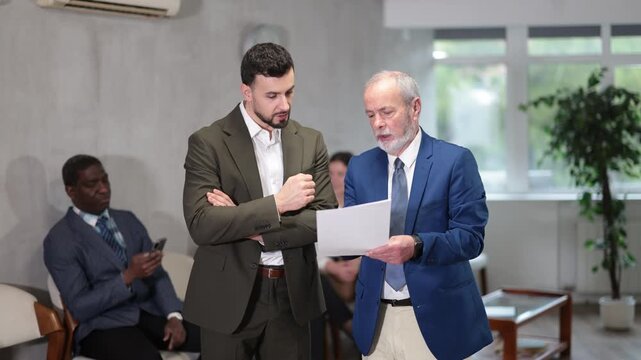  Mature man stands in the corridor against the background of an interview queue and talks to a young businessman. Man gets a job in a company