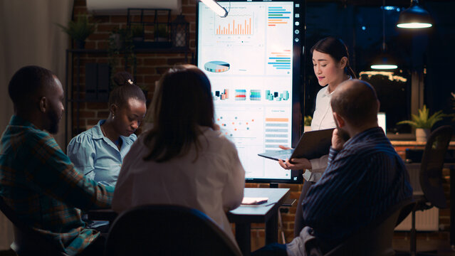 Asian employee presenting company revenue report, showing charts on laptop screen. Research statistics visualization on digital board, diverse team listening to analytics presentation. Tripod shot.