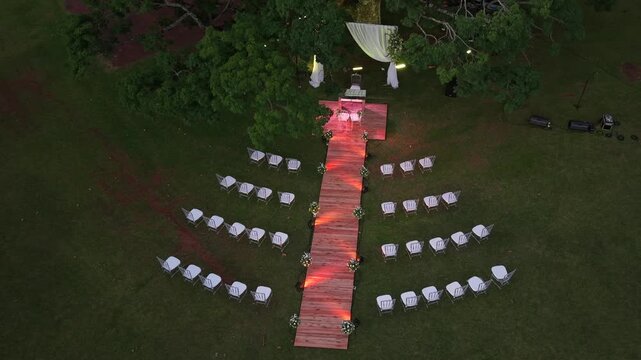 Top down drone descending over outdoor wedding ceremony setup with wooden aisle, white chairs and decorated altar in a garden setting.