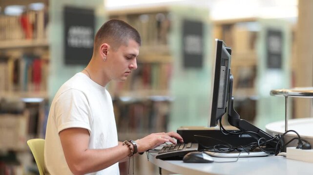European guy is sitting in the library and thoughtfully working at the computer, he is looking for information on the Internet and preparing to write a diploma. High quality 4k footage