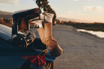 Fototapeta premium Woman wearing casual pants and white sneakers lying upside down in car trunk at sunset, relaxed lifestyle, outdoor scene with road and palm trees in background.