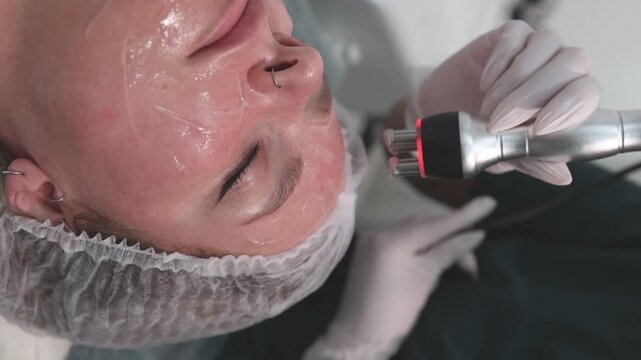 A woman lies relaxed in a clinic, wearing a hairnet, as a specialist uses an RF lifting device on her face. Her skin is coated with a conductive gel. Professional skincare, cosmetic procedure, facial 