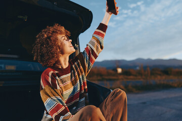 Fototapeta premium Woman smiling outdoors sitting near car trunk wearing rainbow sweater holding smartphone up enjoying lifestyle moment with natural light and clear sky background in casual relaxed pose