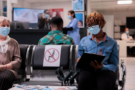 African american patient with mobility problem filling out medical form to undergo tests. Hospital room crowded with diverse people wearing masks to protect themselves from covid.