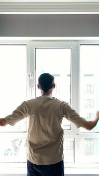 Rear view of a man waking up and opening the curtains in his modern apartment bedroom. He is looking out the window to the bright morning light