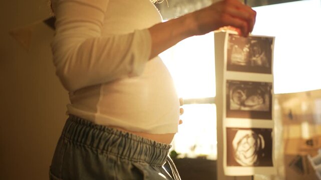 Tender moment of an expecting mother looking at her baby's ultrasound pictures, gently touching her belly while basking in the warm sunlight of her home, anticipating the new arrival