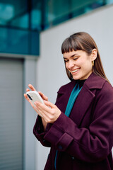 Woman smiling, holding a smartphone, and engaging with digital communication outdoors with an urban background