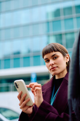 Fototapeta premium Woman standing outdoors, holding and typing on a smartphone, looking at the camera with a calm expression