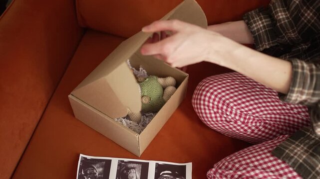 Expectant mother sitting on a couch, opening a gift box with a handmade crochet bunny and a wooden rattle, joyfully examining the baby's ultrasound pictures with heartfelt emotion