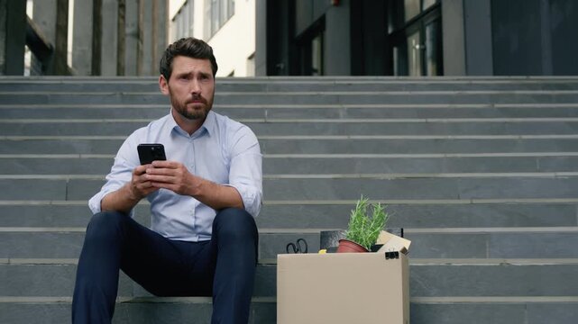 Stressed unemployed Caucasian business man chatting smartphone near office sitting stairs upset fired male businessman dismissal box outside city searching new job online crisis worker problem vacancy