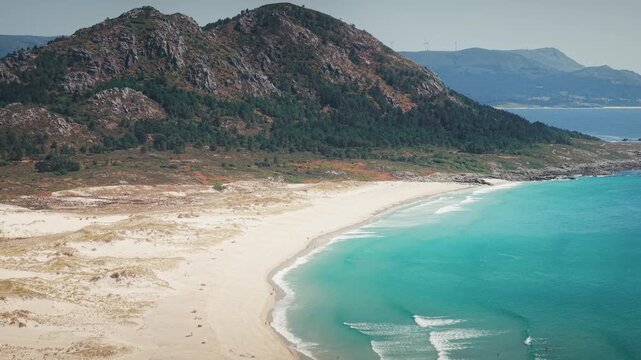 Lateral telephoto drone shot along Monte Louro beach in Galicia, Spain with turquoise water and mountain backdrop.