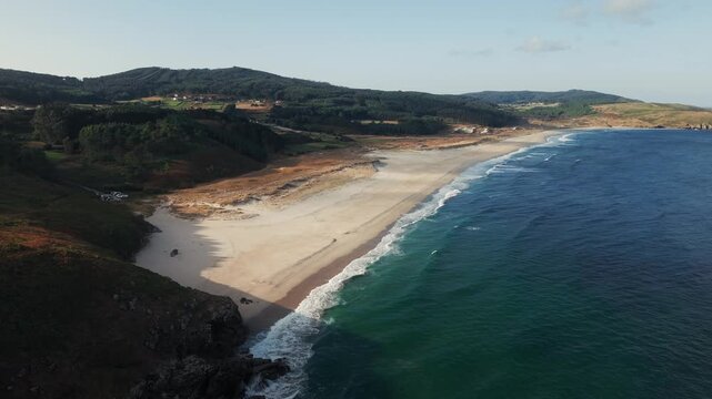 High drone shot pulling back above Praia do Rostro revealing Atlantic waves, sandy coastline and surrounding hills in Galicia, Spain.