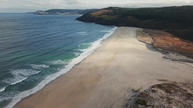 Drone pulling back above Praia do Rostro revealing a vast Atlantic beach with small silhouettes walking along the shore.