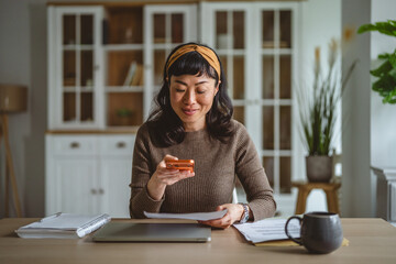 Naklejka na ściany i meble Asian woman managing personal finances with smart phone at home