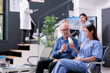Fototapeta premium Physician nurse explaining medication treatment to senior patient while showing painkiller bottle during appointment in hospital reception. Diverse people waiting in lobby before start examination