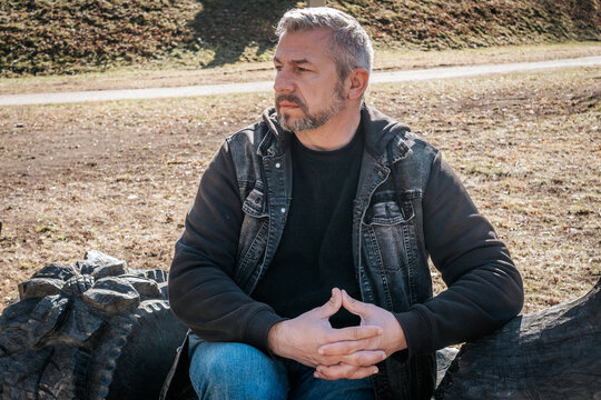 A middle-aged man sits on a carved wooden bench next to a rustic sculpture, looking to the side with a pensive expression in a sunlit park setting. Natural tones and soft shadows enhance the mood.