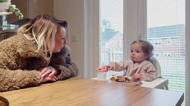 Baby girl and her grandmother clap and smile during dinner at home, captured in a warm indoor family mealtime scene with joyful interaction and bonding
