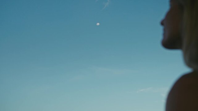 Side profile of a woman looking up at the moon against a clear blue sky. Soft bokeh of her face creates a dreamy, peaceful atmosphere