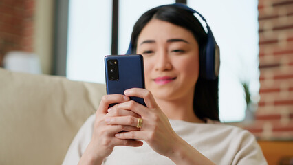 Fototapeta premium Happy young adult person chatting with friend on smartphone while working remotely. Smiling woman sending text messages on mobile phone while listening to music and doing work from home. Handheld shot