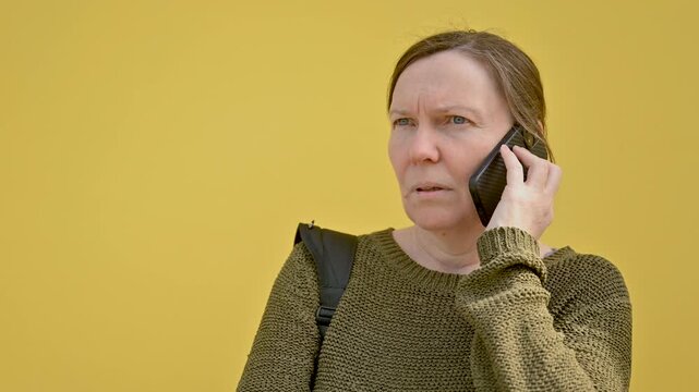 Concerned middle-aged woman talking on smartphone against bright yellow wall, worried expression during phone conversation. 4K.