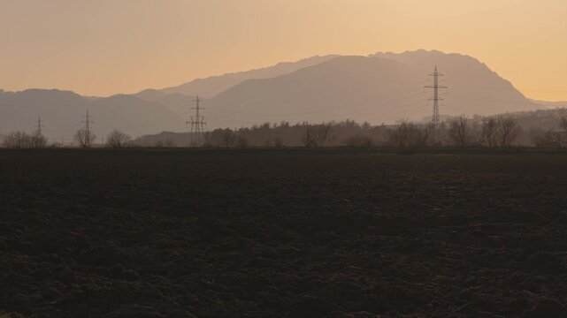 Atmospheric high-voltage power lines towering over plowed fields against a majestic mountain sunset, low altitude lateral tracking drone shot