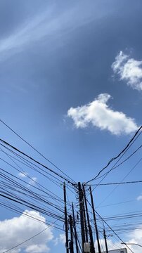 A clear blue sky with scattered white clouds, seen above a complex network of utility poles and tangled electrical wires.