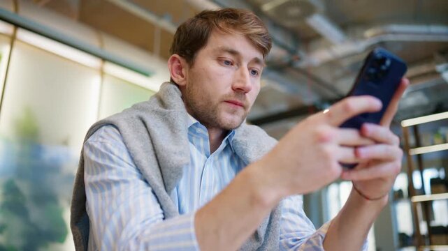 Caucasian man smiling while reading message on smartphone screen. Adult male holding mobile device and looking happy indoors. Sitting at cafe during break time. Positive communication moment.