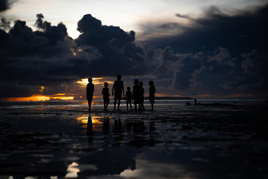View of silhouettes of children stand along the shore as the sun sets, casting a golden glow against the dark clouds, Lomlom, Reef, Solomon Islands.