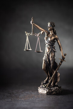A bronze statue of Justice stands against a dark, textured background with dramatic side lighting. The figure holds scales symbolizing justice in one hand and a sword in the other