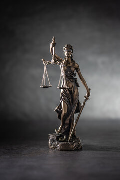 A bronze statue of Justice stands against a dark, textured background with dramatic side lighting. The figure holds scales symbolizing justice in one hand and a sword in the other