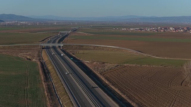Striking modern highway and overpass with moving traffic in a picturesque rural landscape at golden hour near Brasov Romania, low altitude aerial forward tracking shot