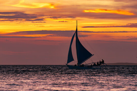 View of a lone sailboat glides across the rippling water against a backdrop of a fiery sunset painting the sky with orange and purple hues, Malay, Philippines.