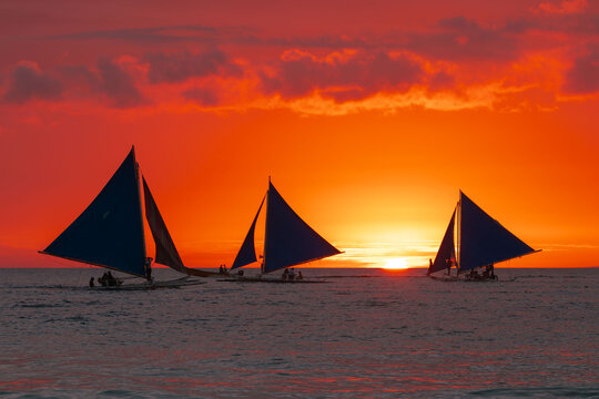 View of silhouettes of sailboats gliding across the water against a fiery sunset, their sails catching the last light of day in Boracay, Malay, Philippines.