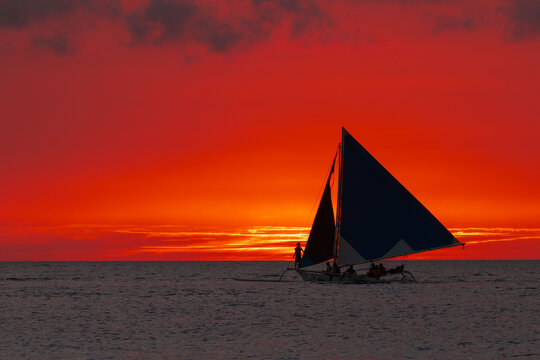 View of a sailboat silhouetted against a fiery sunset sky, its sails catching the last light on the horizon, Boracay, Western Visayas, Philippines.