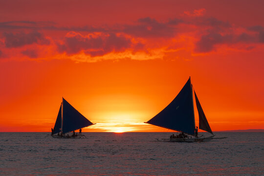View of vibrant orange sunset casting silhouettes of sailboats gliding across the serene sea, painting a tropical dream, Boracay, Western Visayas, Philippines.