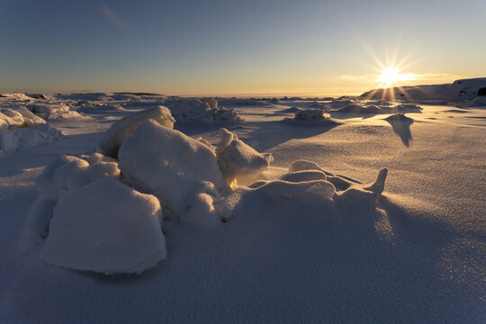 View of a glistening, snow-covered landscape dotted with icy formations reflecting the warm glow of the setting sun, Kandalaksha, Murmansk Oblast, Russia.
