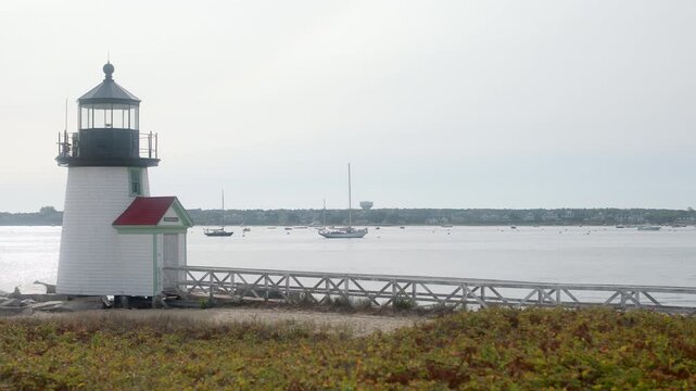 Cinematic gimbal truck left at Brant Point Lighthouse in Nantucket, Massachusetts, revealing sailboats anchored in the calm harbor under soft coastal daylight.