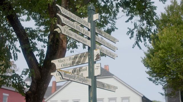 Handheld shot of a weathered destination signpost in Nantucket, Massachusetts, pointing to local landmarks beneath leafy trees in soft daylight.