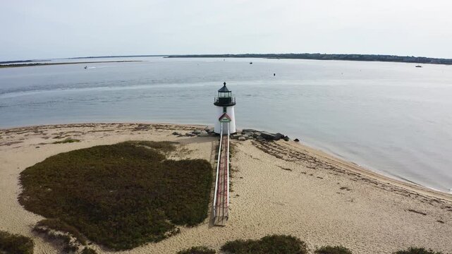 Cinematic drone fly-out from Brant Point Lighthouse in Nantucket, Massachusetts, revealing sandy shoreline and expansive harbor waters from above.