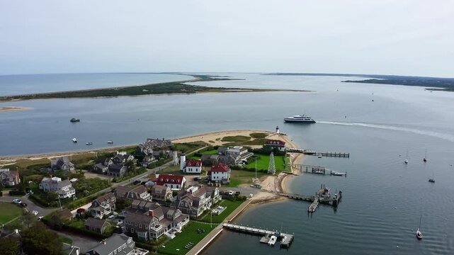 A drone flies over Brant Point Lighthouse in Nantucket, Massachusetts, as a ferry glides across the harbor, revealing the peninsula, coastal homes, and layered shoreline from above.