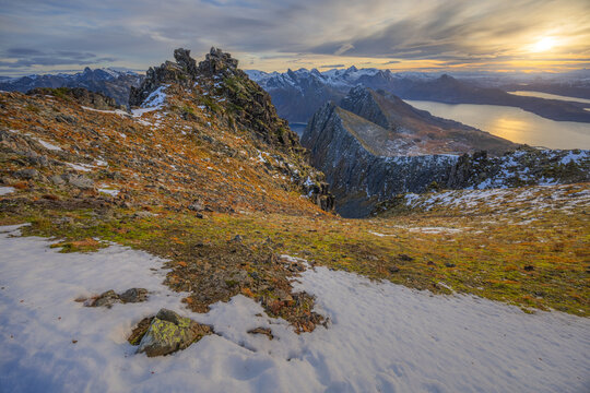 View of rugged peaks dusted with snow rise above the golden autumn tundra, overlooking the serene fjord reflecting the sunset, Senjahopen, Troms, Norway.