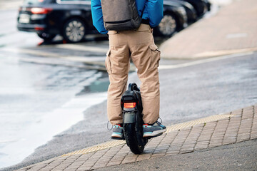 Commuter on an electric unicycle balancing on brick sidewalk with wet asphalt, back view. Person riding electric unicycle with red tail light on paved street wearing tan cargo pants and blue jacket © Tricky Shark