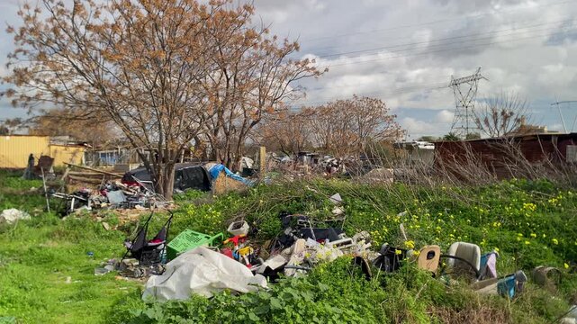 Accumulations of illegally dumped household waste and trash polluting a grassy urban area with sparse trees and scattered makeshift shelters in Seville