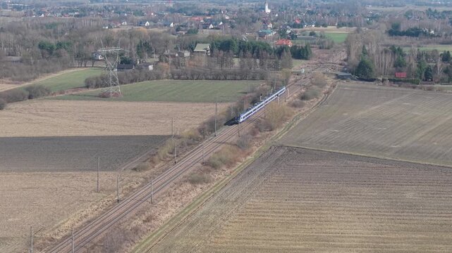 Aerial drone footage of a modern passenger train travelling through a flat rural landscape with agricultural fields and bare winter trees. Electric railway with overhead power lines cuts diagonally ac
