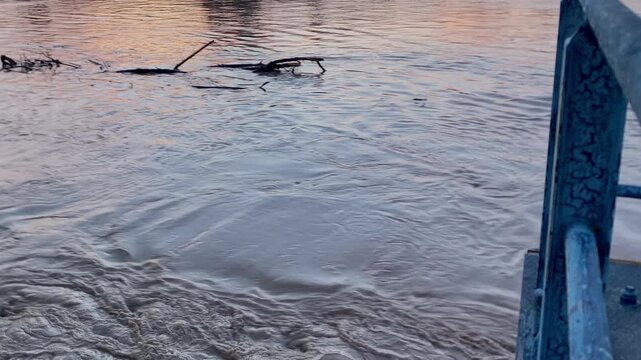 Muddy water flowing in a river, showing high levels and erosion from recent heavy rainfall. A blue railing overlooks the scene