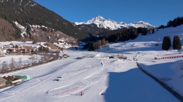 Aerial view of cross country ski trail in Savognin, Switzerland with snowy alpine landscape, mountains and person with kid on sleigh enjoying a sunny winter day. Flyover shot.