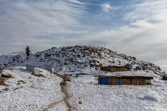 View of a snowy hill dotted with small wooden structures under a lightly clouded sky, contrasting the cold white with the earthy wood, Rawalakot, Pakistan.