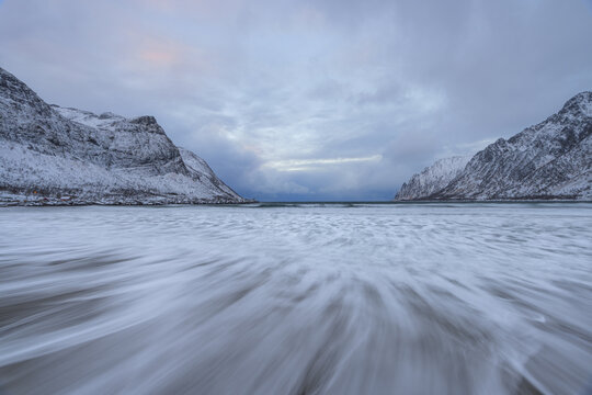 View of the beach where the tide blurs the line between the shore and the sea, framed by snow-covered mountains under a pale, overcast sky, Senjahopen, Troms, Norway.