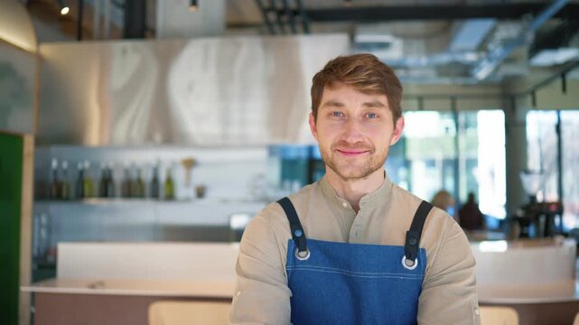 Male cafe worker smiling at camera in restaurant interior. Man wearing apron standing near bar counter. Hospitality service. Employee welcoming visitors inside modern cafe with open kitchen behind.